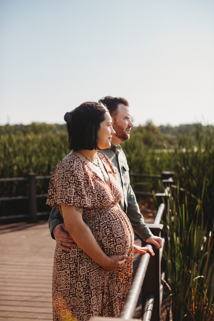 Pregnant mom and husband stand side by side overlooking Reeds Lake. 