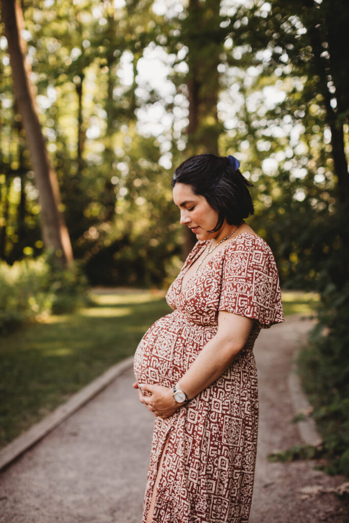 Pregnant woman holds baby bump on wooded path by Reeds Lake for maternity photos