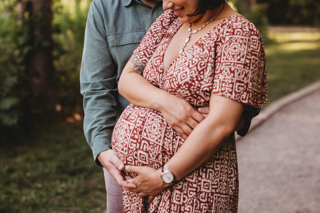 Couple wraps their arms around pregnant mom's baby bump while posing for maternity photo in wooded area by Reeds Lake